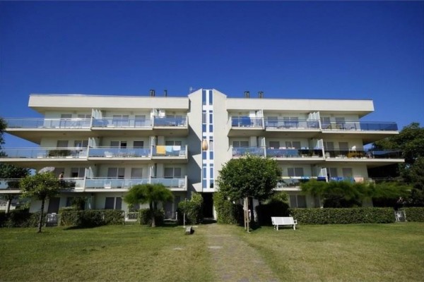 Čtyřpatrová obytná budova s balkony a zahradou před ní na pozadí modré oblohy.