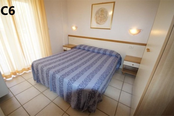 Ložnice s dvojlůžkem, bočními stolky a světle modrým přehozem, vedle okna s jemnými závěsy.  