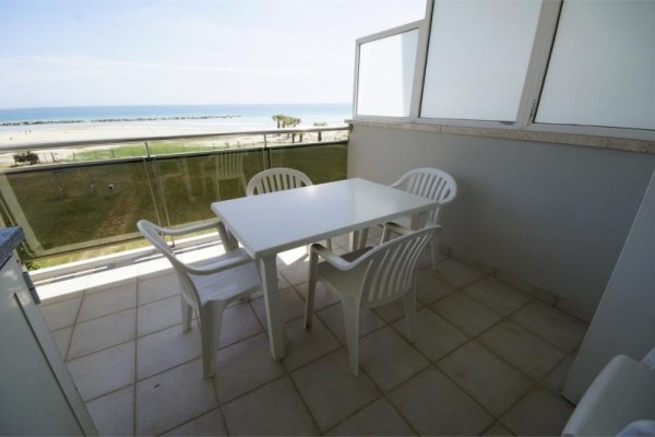 Balkon s výhledem na pláž a moře, vybavený čtyřmi bílými plastovými židlemi a stolem.