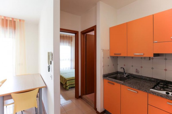 Moderní kuchyně s oranžovými skříňkami vedle jídelního koutu a dveří do ložnice.