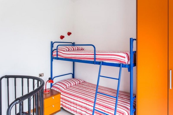 Palanda s červeno-bílým proužkovaným povlečením, vedle je oranžová skříň a noční stolek s lampou.