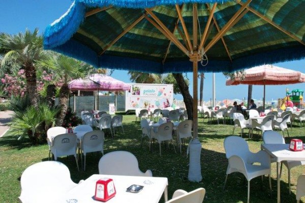 Restaurace s venkovním posezením se slunečníky a palmami na pozadí.  