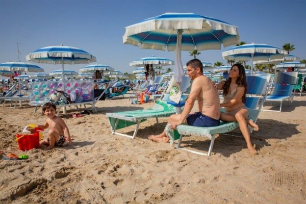 Rodina relaxuje na pláži pod modrými slunečníky, dítě si hraje v písku.