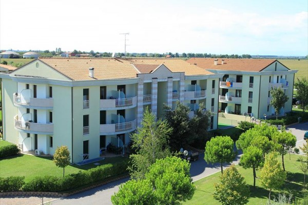 Třípatrová bytová budova s balkony obklopená zelení a stromy.
