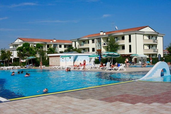 Venkovní bazén s lidmi u několika bílých budov a slunečníky kolem něj.