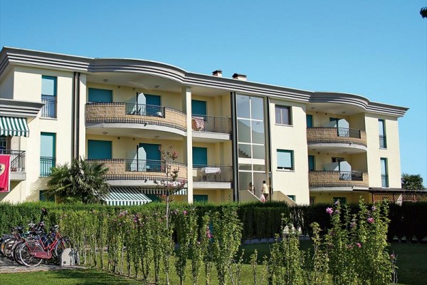 Třípatrová moderní budova s balkony a zelenými okenními roletami obklopená zahradou a zaparkovanými koly.