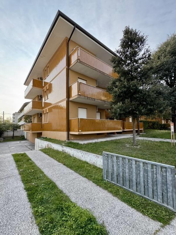 Moderní bytový dům se třemi patry a balkony, obklopen zelení a stromem.