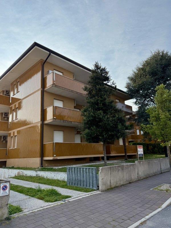 Třípatrový bytový dům s balkóny obklopený stromy.