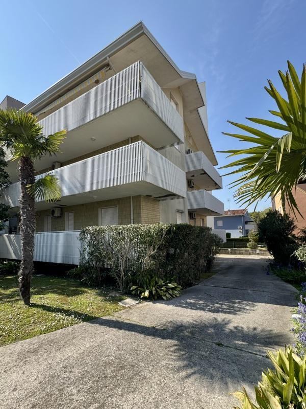 Moderní třípatrová budova s balkonem obklopená zelení a palmami.
