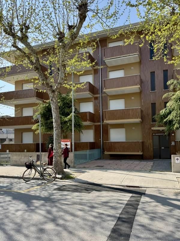 Vícepatrová budova s balkony a stromem v popředí.