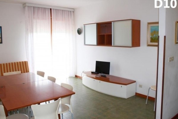 Jednoduše zařízený obývací pokoj s televizí, stolem a židlemi u velkého okna s bílými závěsy.