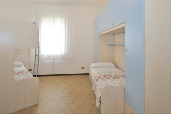 Pokoj s patrovou a samostatnou postelí, světlým nábytkem a oknem se záclonou.
