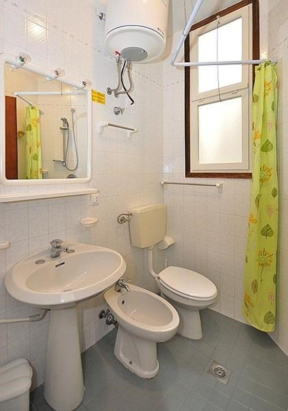 Koupelna s keramickým umyvadlem, bidetem a záchodem, sprcha za zeleným závěsem a ohřívač vody na stěně.